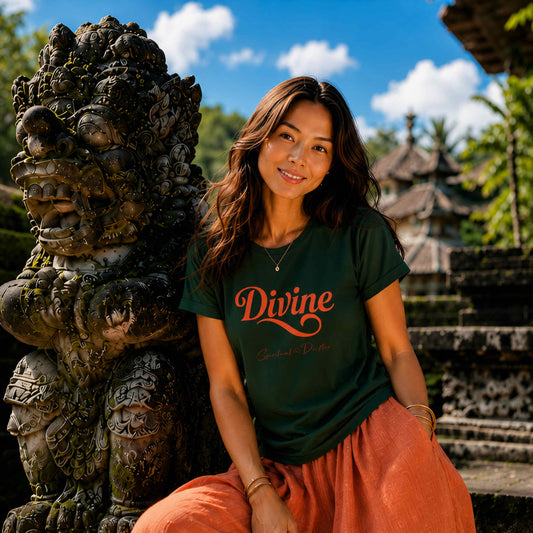 Woman sitting in a temple with intricate carvings in Bali Indonesia. She is wearing orange silk pants and a Spiritual Drifter forest green shirt with the graphic "Divine" on the t shirt