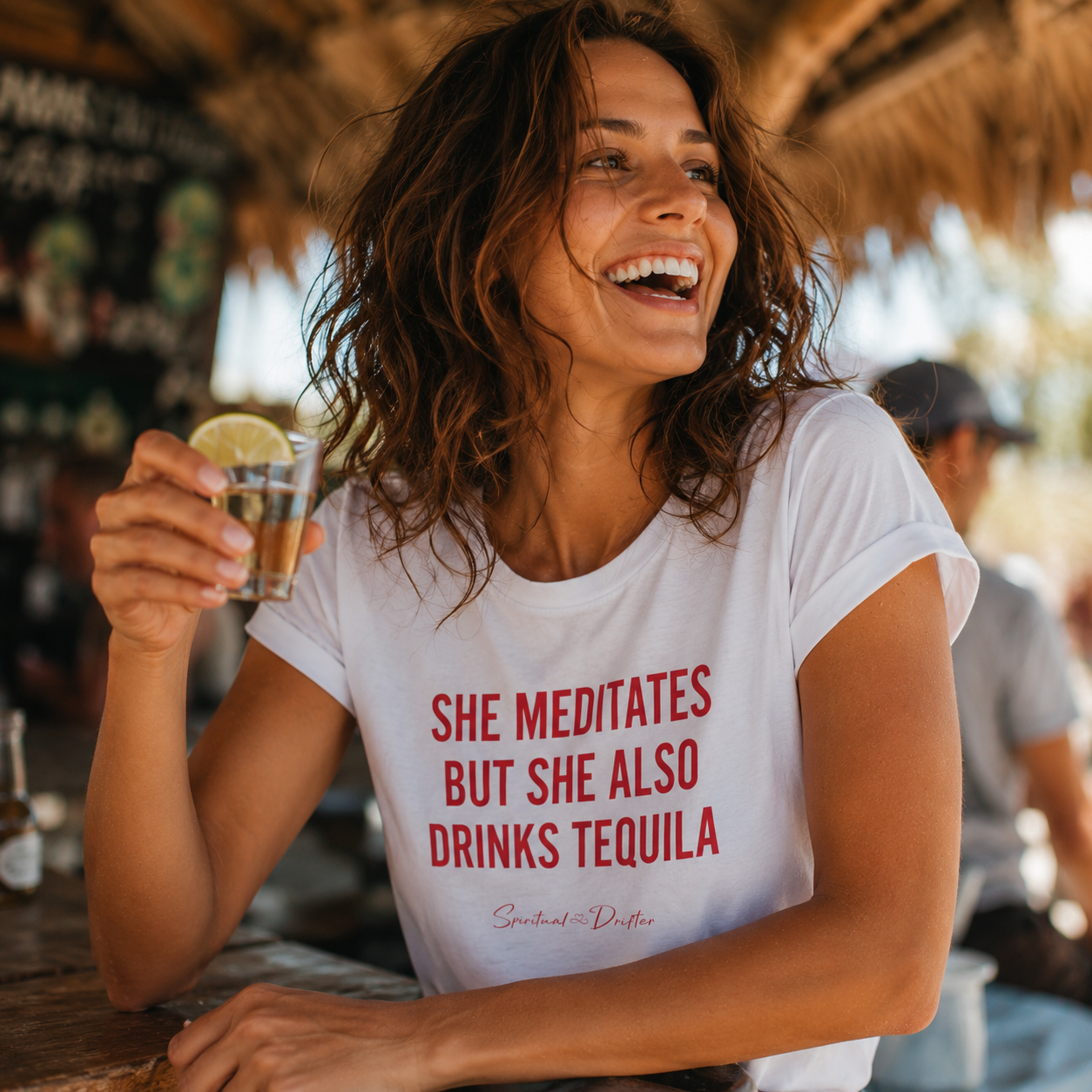 Woman in a white t-shirt with humorous text holding a tequila shot in a tropical setting. Wearing Spiritual Drifter t-shirt with red text 'SHE MEDITATES BUT SHE ALSO DRINKS TEQUILA' on a retreat in Cancun Mexico. Women's spiritual graphic tee. Relaxed beach lifestyle fashion for meditation lovers, yoga enthusiasts, wellness travelers, and free-spirited women. Meditation and yoga clothing for awakened souls.