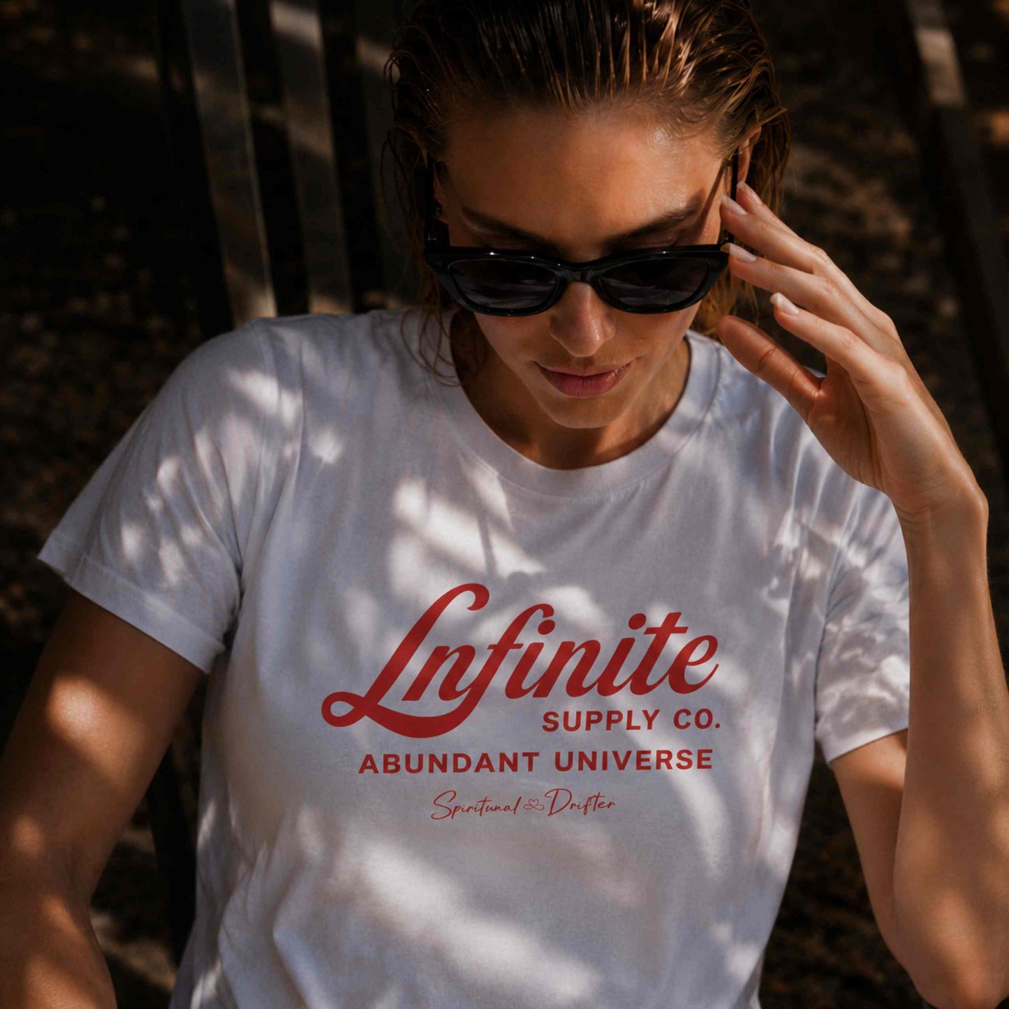 Woman wearing Spiritual Drifter ‘Infinite Supply Abundant Universe’ graphic cotton t-shirt sitting in the shade by a pool. Elevated spiritual apparel Inspired by Abraham Hicks and the Law of Attraction. 