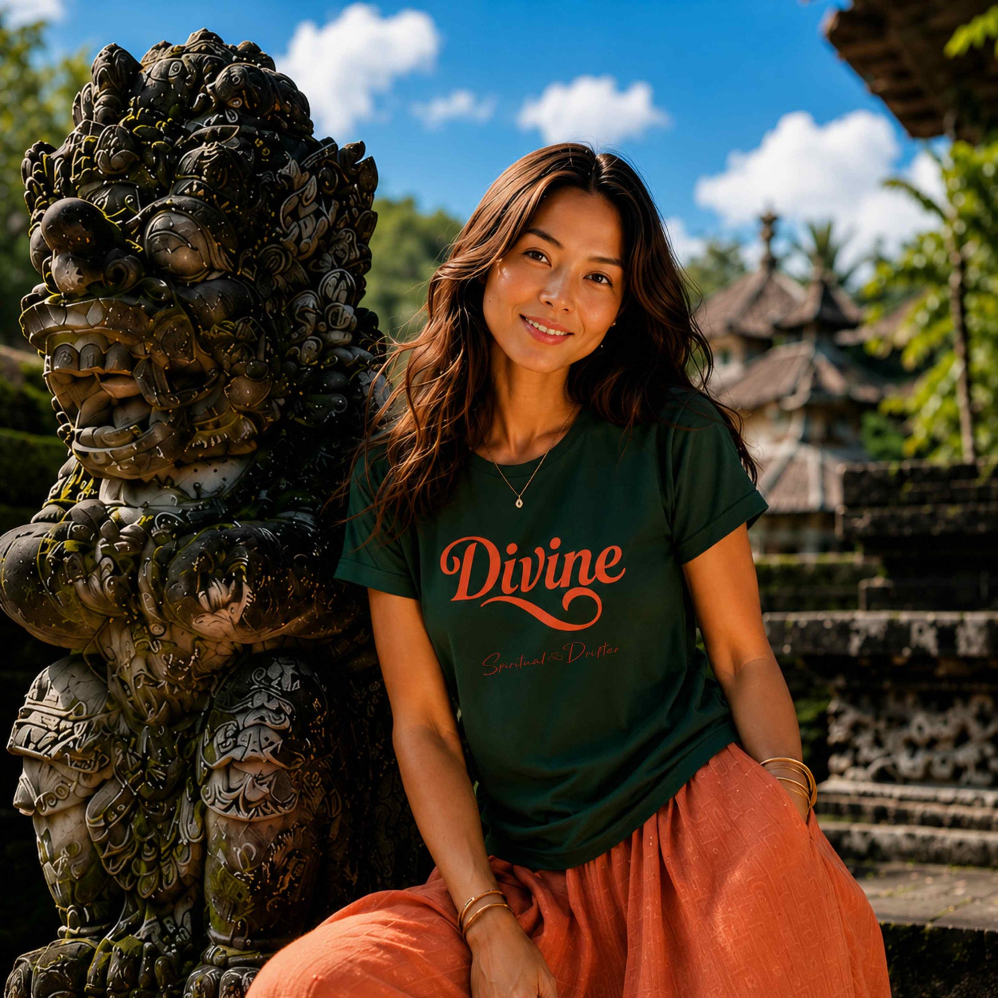 Woman sitting in a temple with intricate carvings in Bali Indonesia. She is wearing orange silk pants and a Spiritual Drifter forest green shirt with the graphic "Divine" on the t shirt