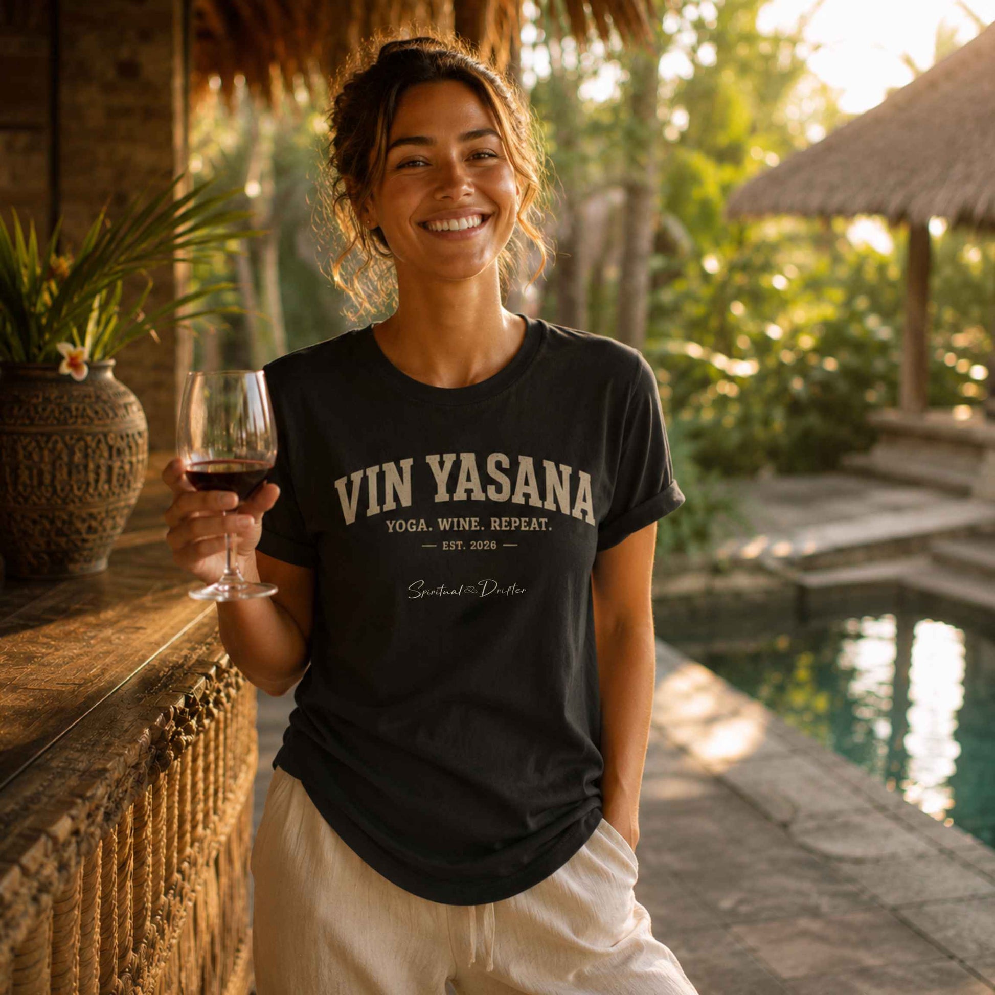 Woman holding a glass of wine wearing a Spiritual Drifter 'VIN YASANA' t-shirt by a poolside in Bali. Yoga and elevated consciousness apparel.