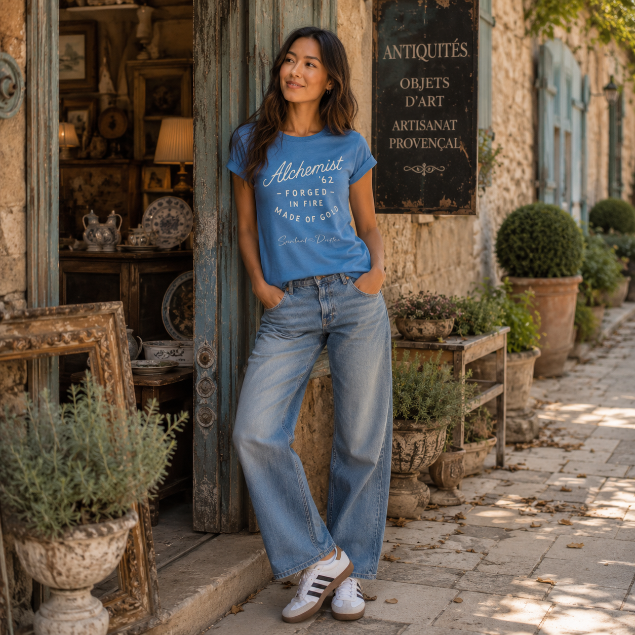 Woman wearing a blue Spiritual Drifter "Alchemist, forged in fire" t-shirt and jeans standing in front of an elegant antique shop in Provence, France.