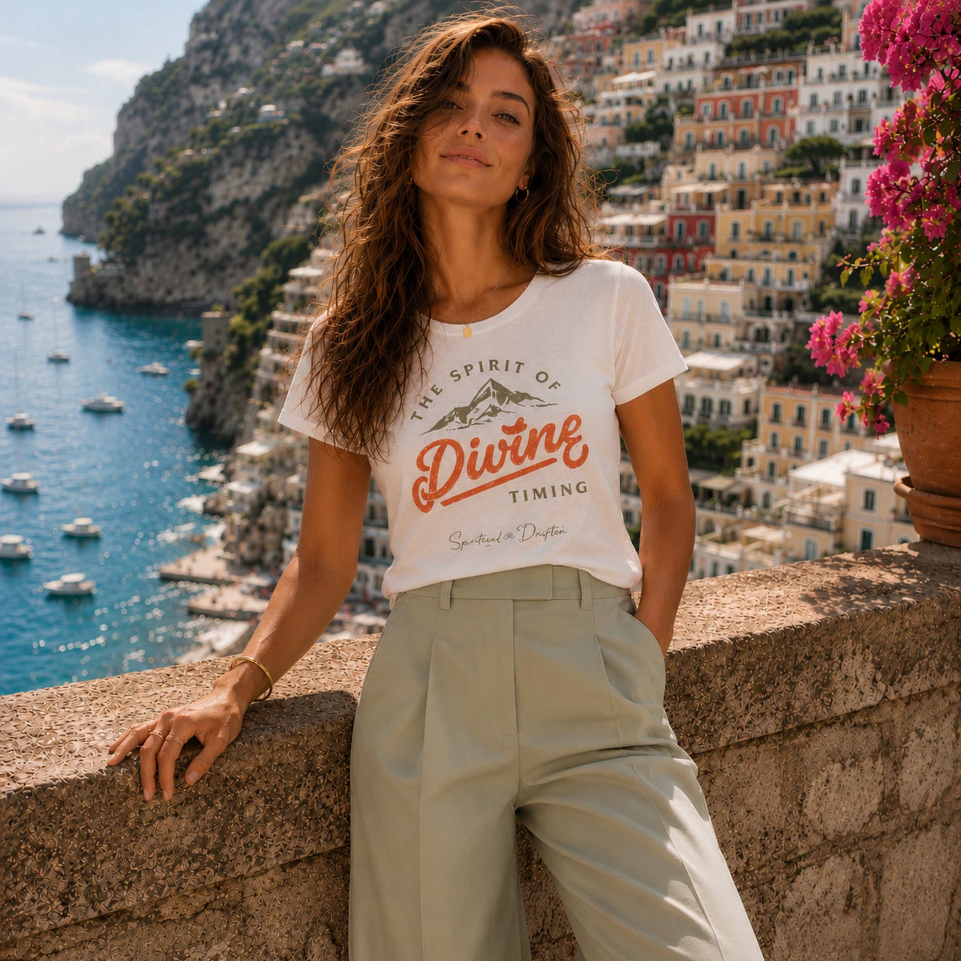 Woman wearing a t-shirt with 'The Spirit of Divine Timing' design, standing in front of Positano in Italy. Spiritual Drifter elevated conscious living apparel. Cotton curved hem cap sleeved woman's shirt.